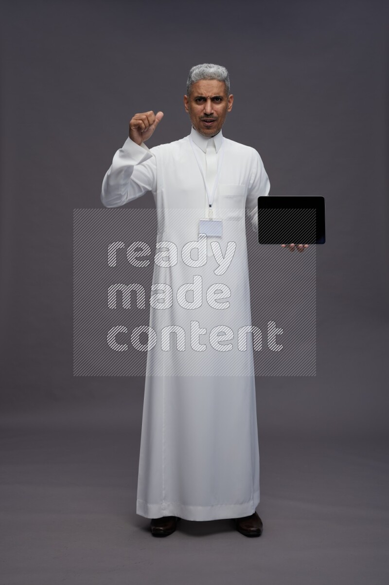 Saudi man wearing thob with neck strap employee badge standing showing tablet to camera on gray background
