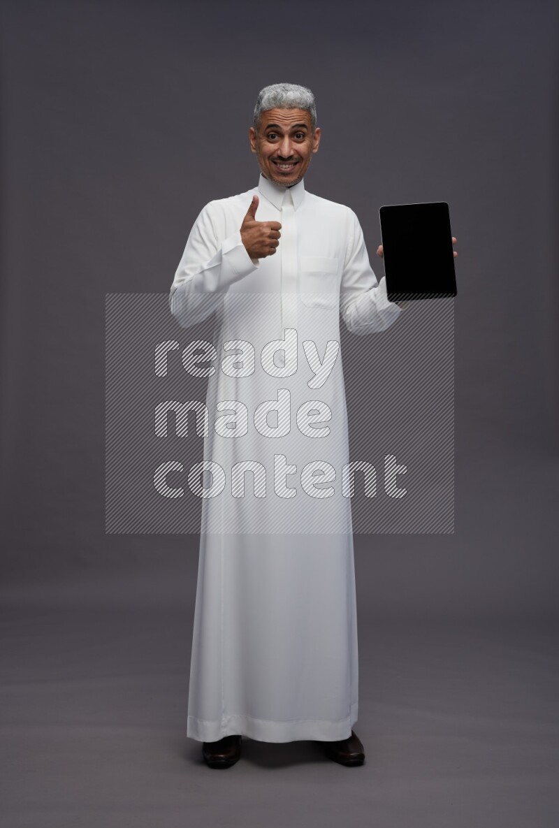 Saudi man wearing thob standing showing tablet to camera on gray background