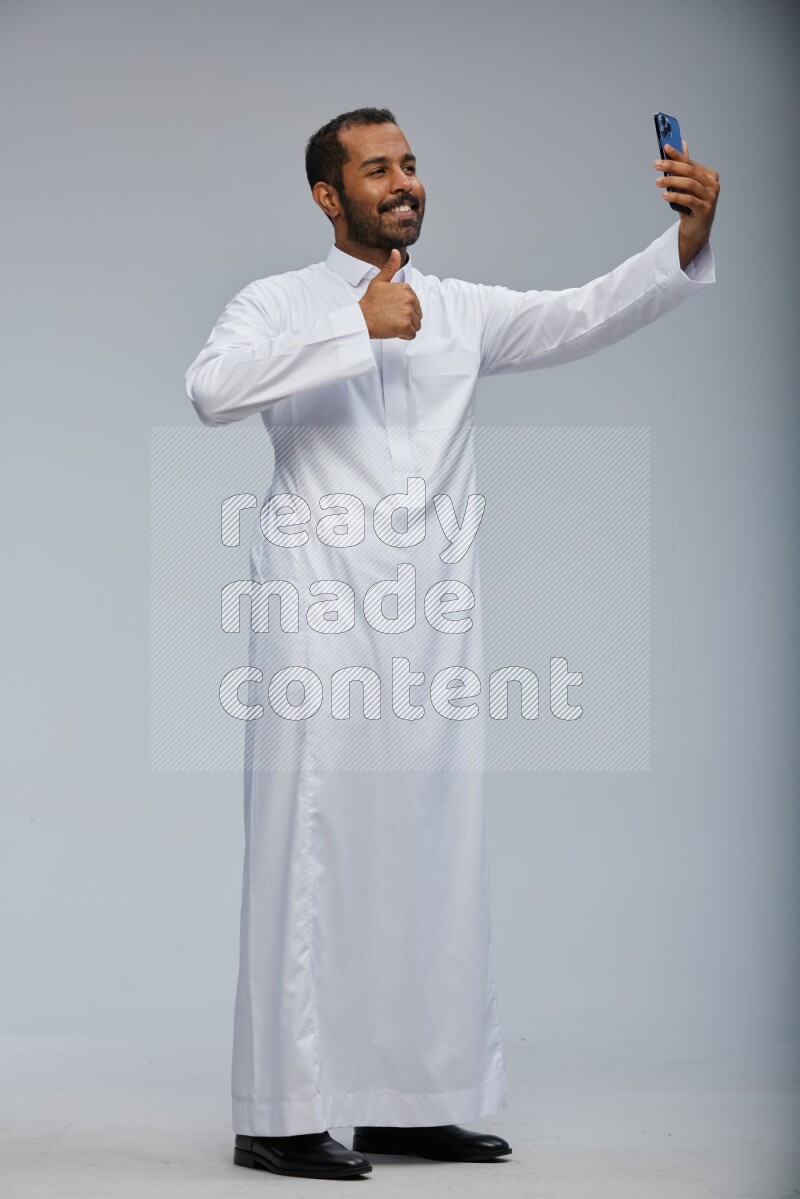 Saudi man wearing Thob standing taking selfie on Gray background