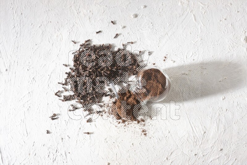 وعاء زجاجي مقلوب ممتلئ ببودرة القرنفل وتناثرت البودرة منه مع حبوب القرنفل المنتشرة على أرضية بيضاء