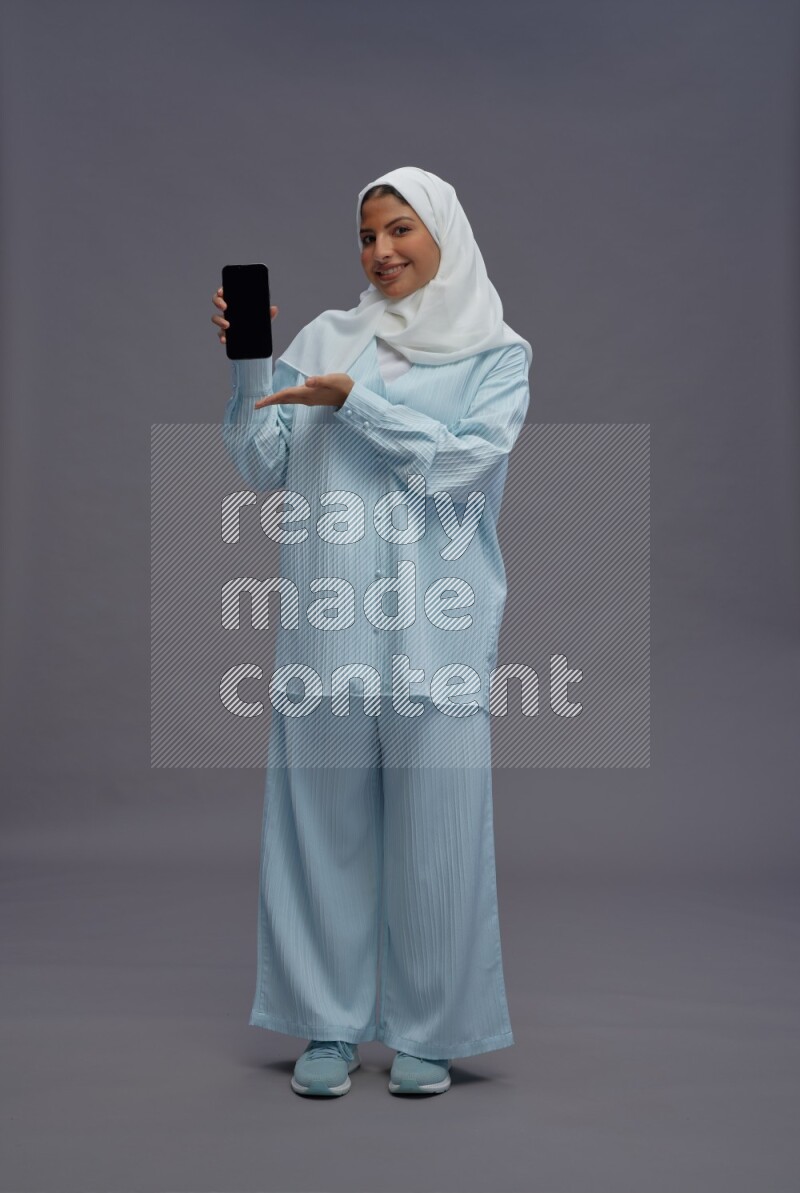 Saudi woman wearing hijab clothes standing showing phone to camera on gray background