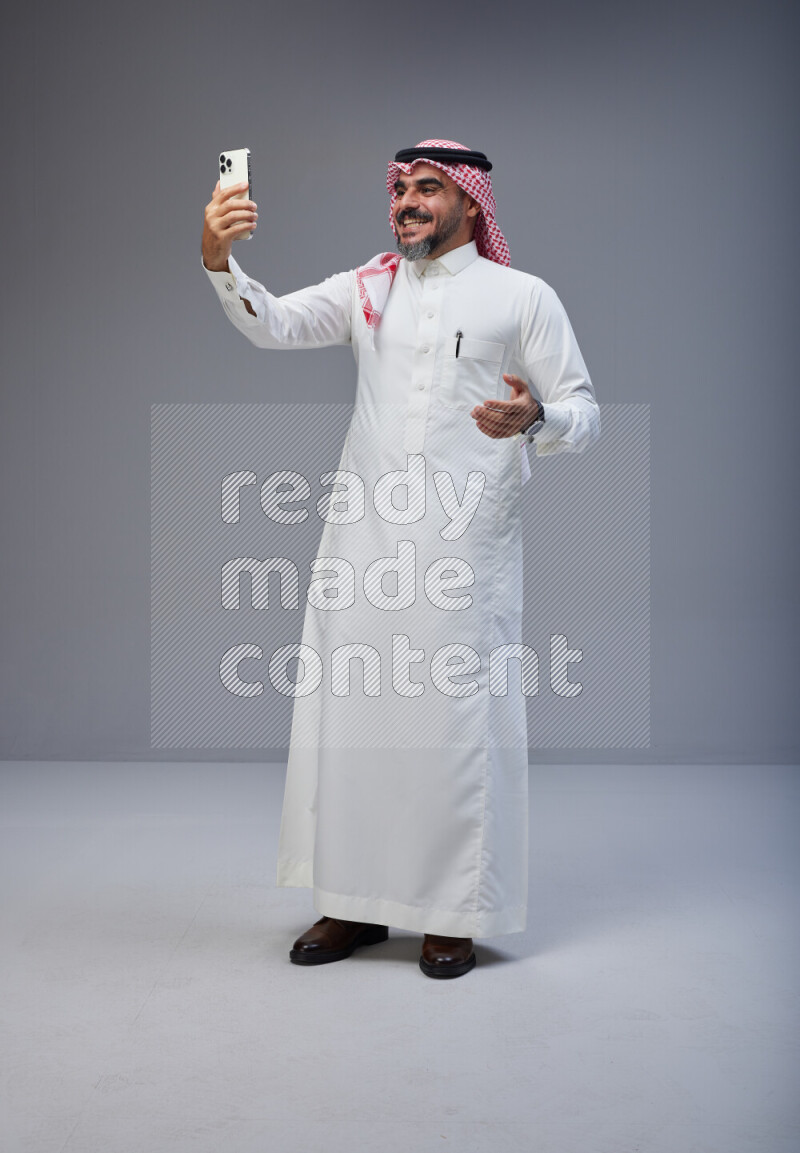 Saudi man Wearing Thob and red Shomag standing taking selfie on Gray background