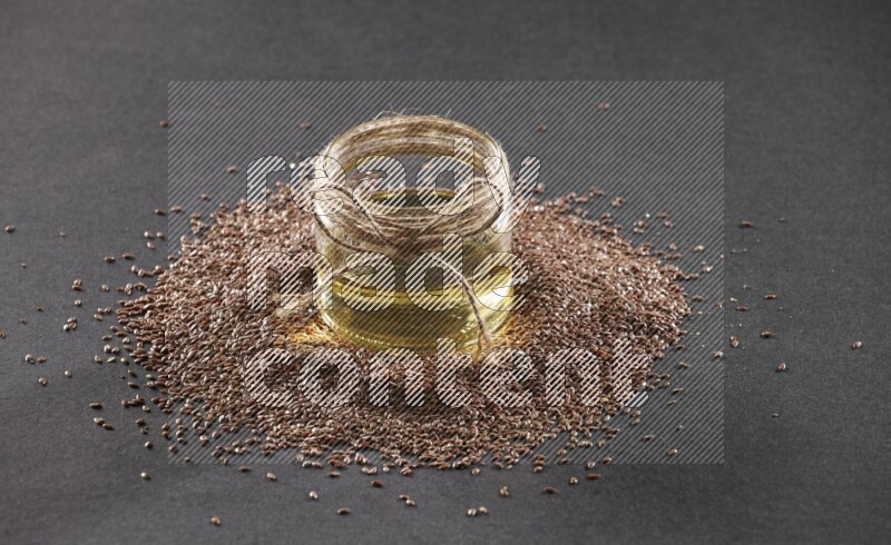 A glass jar full of flaxseeds oil surrounded by the seeds on a black flooring