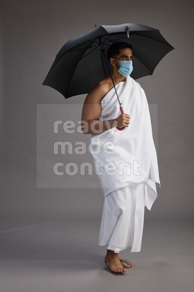 A man wearing Ehram and mask Standing holding umbrella on gray background on gray background
