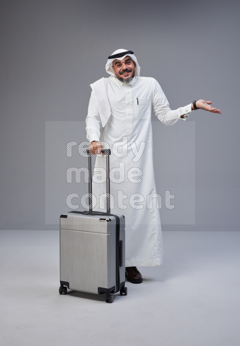 Saudi man wearing Thob and white Shomag standing holding Travel bag on Gray background