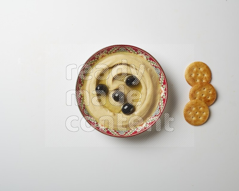 Hummus in a red plate with patterns garnished with black olives on a white background