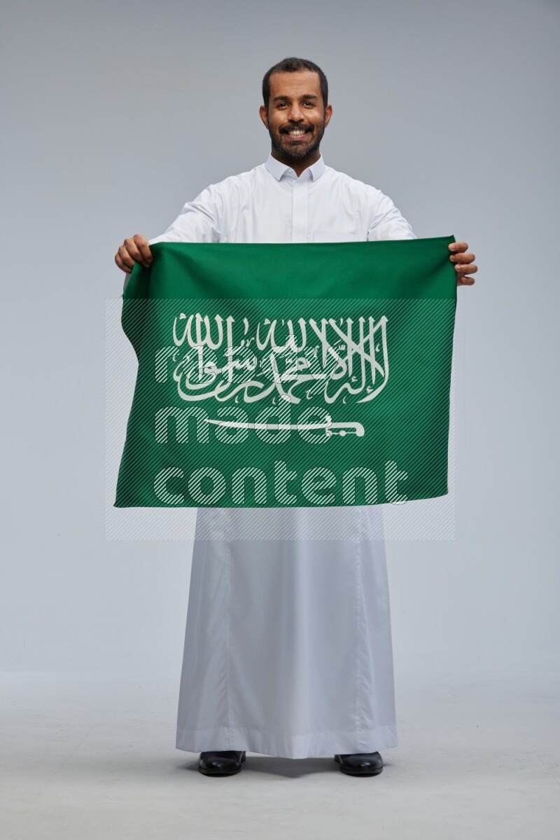 Saudi man wearing Thob standing holding Saudi flag on Gray background