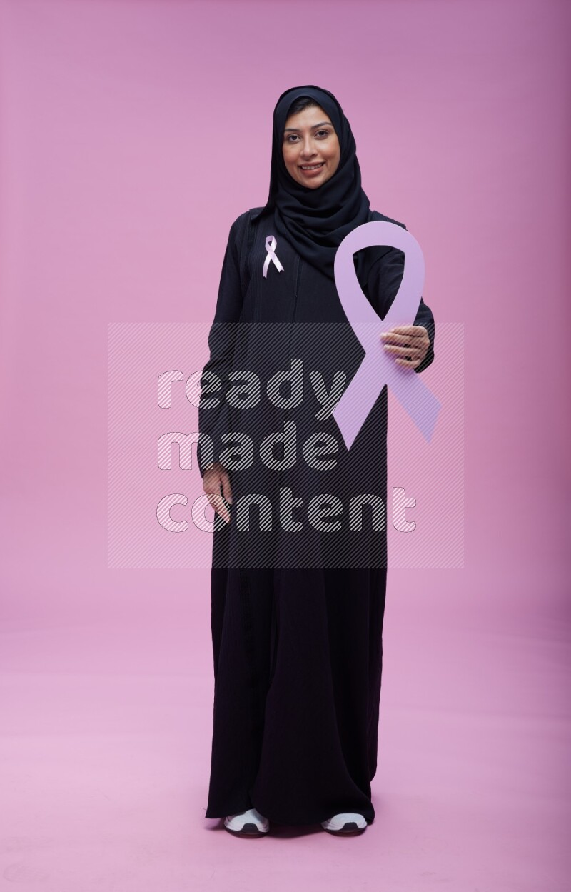 Saudi woman wearing pink ribbon on abaya standing holding awareness ribbon on plain pink background