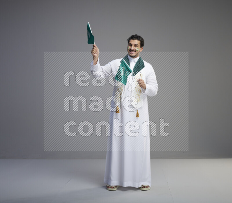 A Saudi man standing wearing thob and saudi flag scarf with face painting holding small Saudi flag on gray background