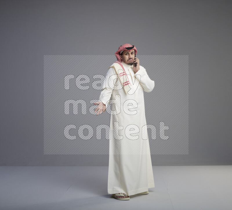 A Saudi man standing wearing thob and red shomag talking on phone on gray background