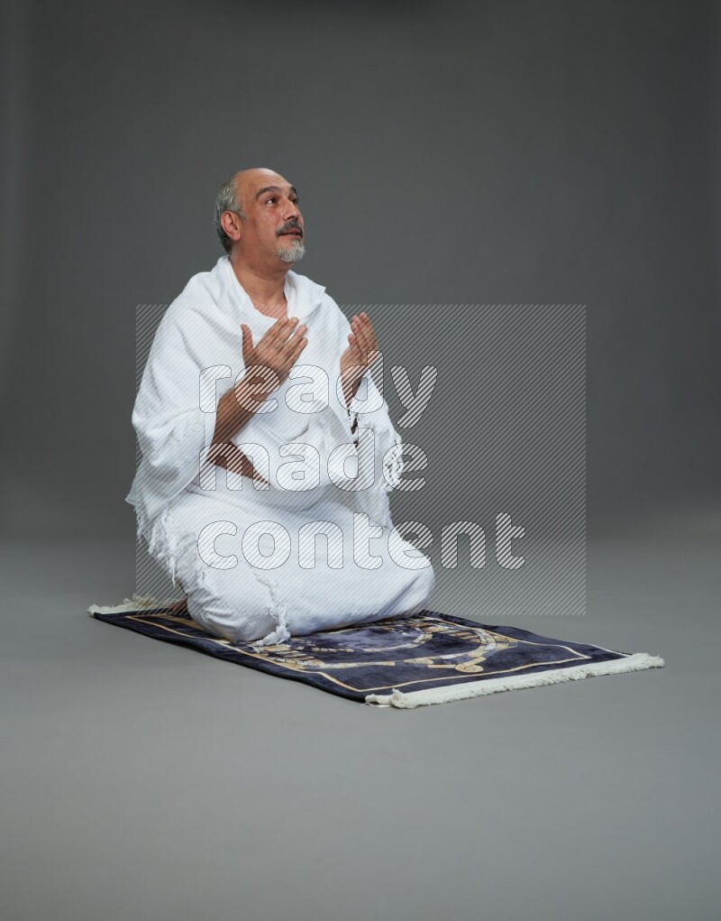 A man wearing Ehram sitting on prayer mat dua'a on gray background