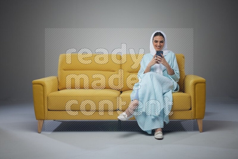 A Saudi woman wearing a light blue Abaya and white head scarf sitting on a yellow sofa and texting on her phone on a grey background
