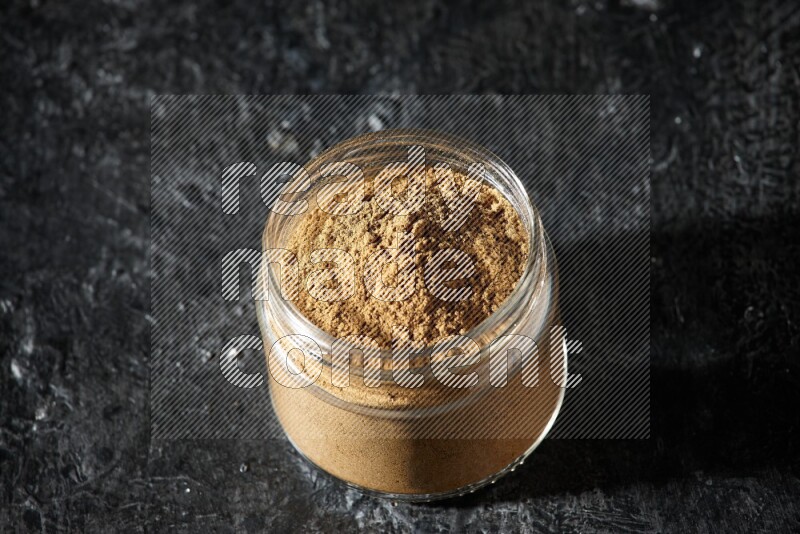A glass jar full of allspice powder on a textured black flooring