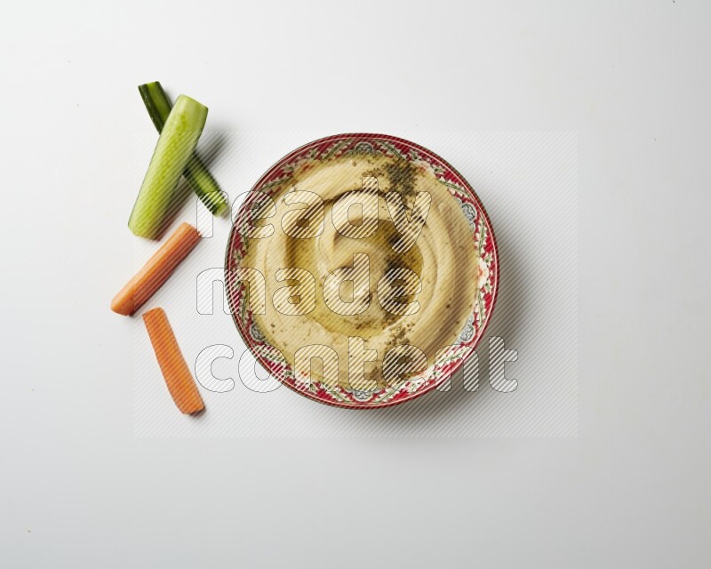Hummus in a red plate with patterns garnished with zaatar & sumak on a white background
