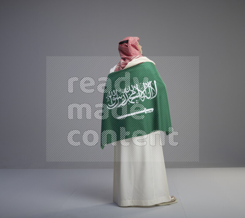 A Saudi man standing wearing thob and red shomag wrapping big saudi flag on gray background