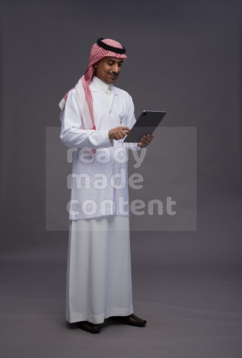 Saudi man wearing thob with lab coat and shomag with pocket employee badge standing working on tablet on gray background