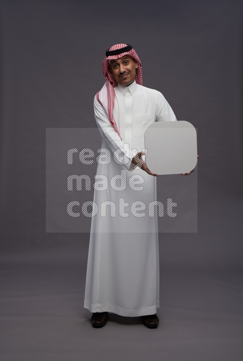 Saudi man wearing thob and shomag standing holding social media sign on gray background