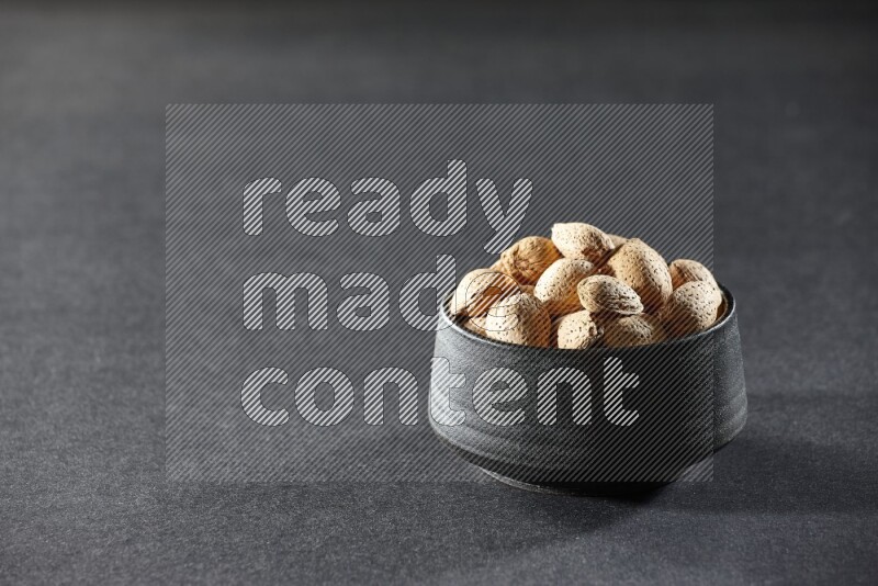 A black pottery bowl full of almonds on a black background in different angles