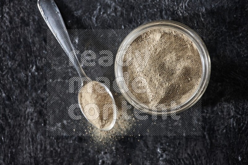 A glass jar and a metal spoon full of white pepper powder on textured black flooring