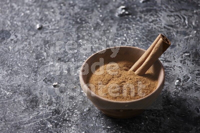 Wooden bowl full of cinnamon powder and a cinnamon stick on a textured black background