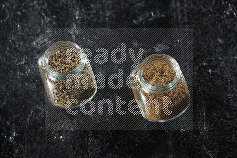 2 glass spice jars full of cumin powder and seeds on a textured black flooring