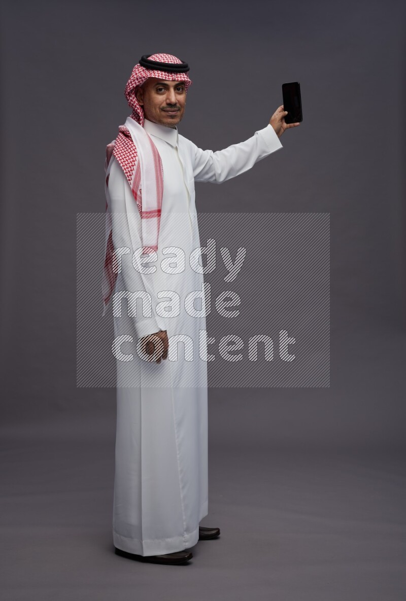 Saudi man wearing thob and shomag standing taking selfie on gray background