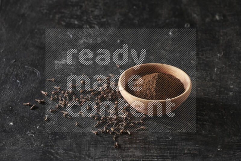 A wooden bowl full of cloves powder on a textured black flooring