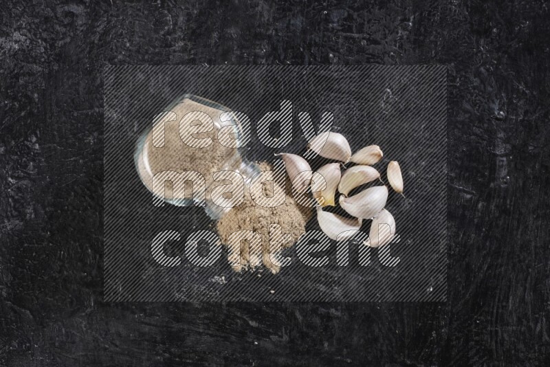 A glass spice jar full of garlic powder flipped over with the powder came out on a textured black flooring