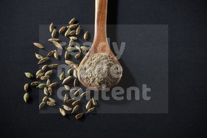 A wooden ladle full of cardamom powder and cardamom seeds beside it on black flooring