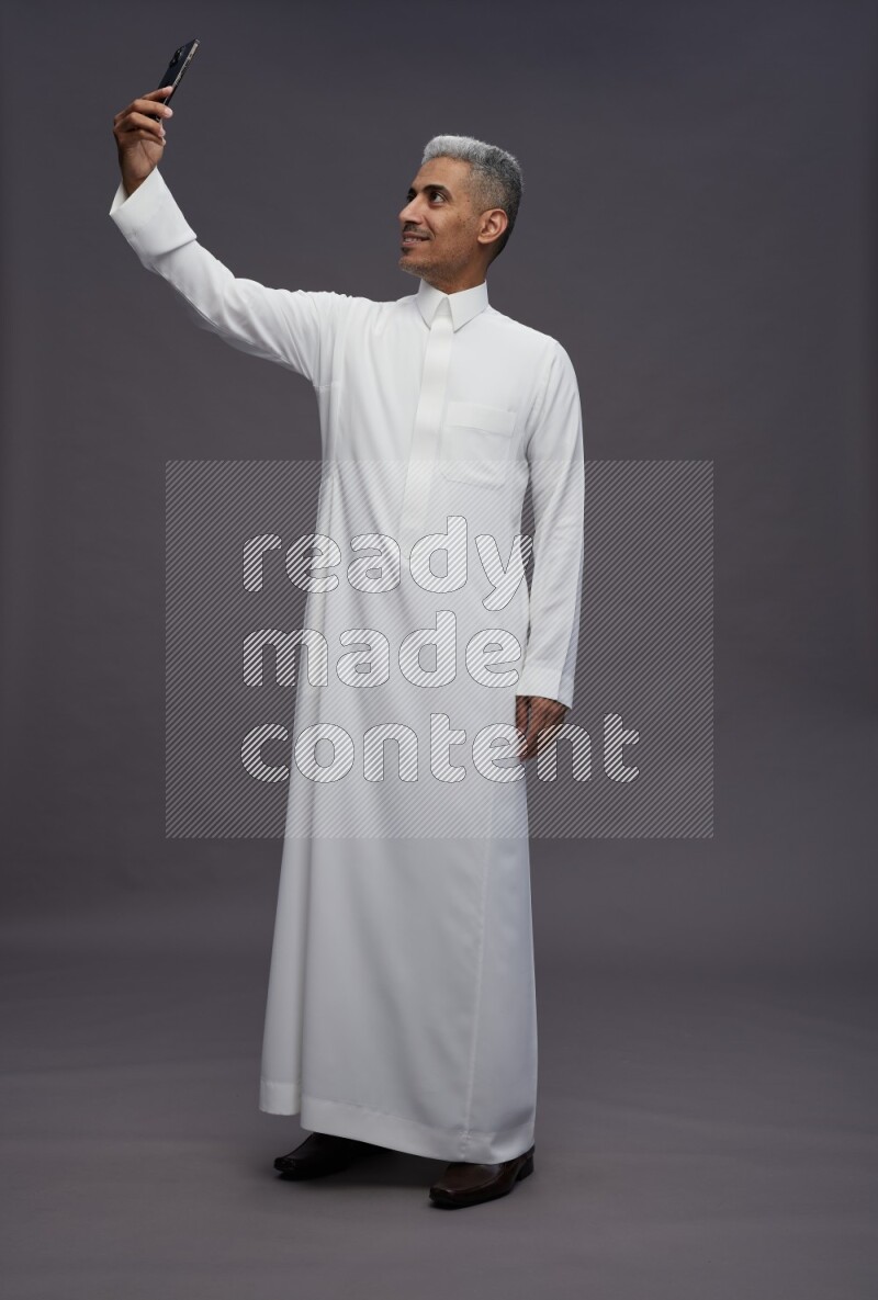 Saudi man wearing thob standing taking selfie on gray background
