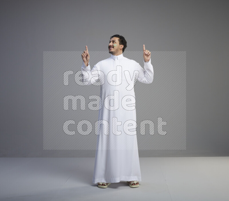 A Saudi man standing wearing thob with face painting interacting with the camera on gray background