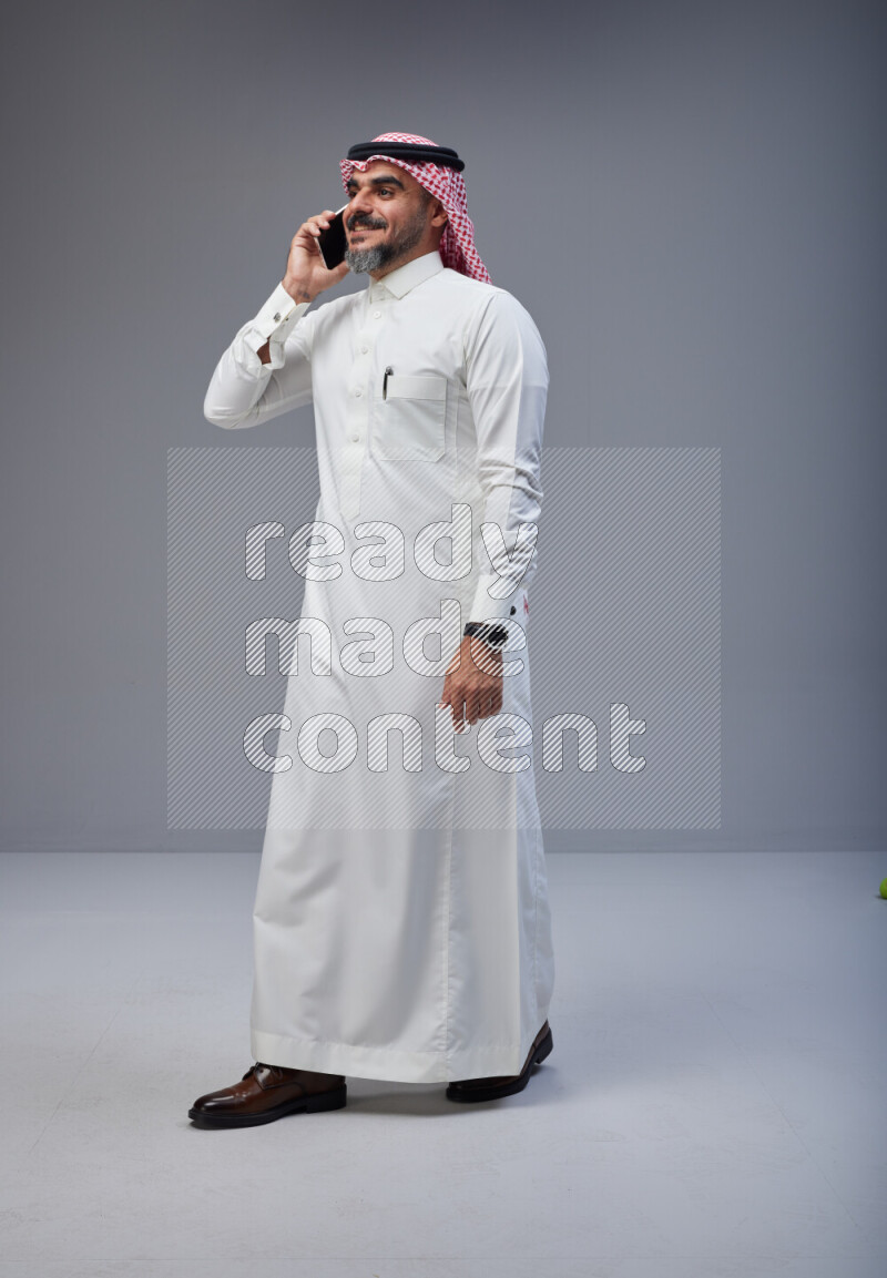 Saudi man Wearing Thob and red Shomag standing talking on phone on Gray background