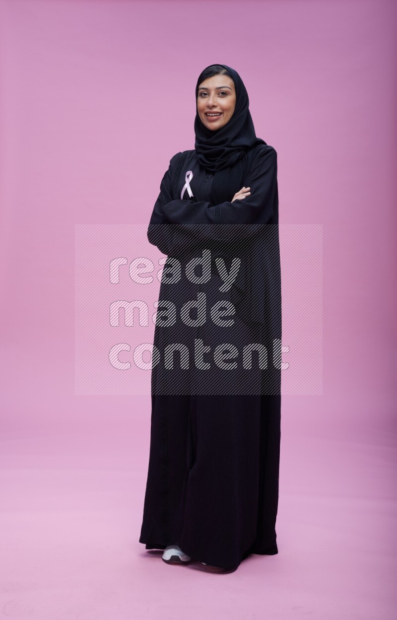 Saudi woman wearing pink ribbon on Abaya standing crossed arms on pink background