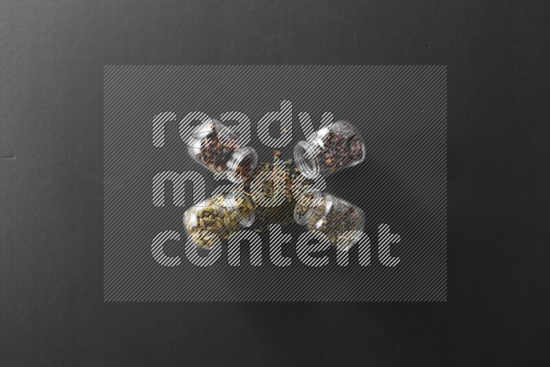 4 glass spice jars full of cloves, cardamom, basil and star anise on black flooring