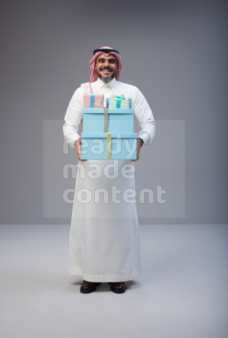Saudi man Wearing Thob and red Shomag standing holding gift box on Gray background