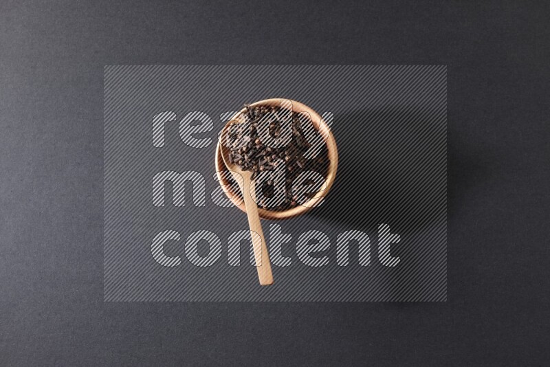 A wooden bowl and a wooden spoon full of cloves on a black flooring