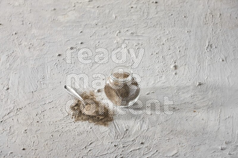 A glass spice jar full of black pepper powder and metal spoon full of it on textured white flooring