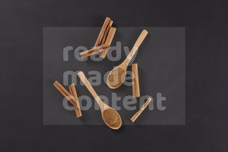 Two wooden spoons full of cinnamon powder with cinnamon sticks on black background