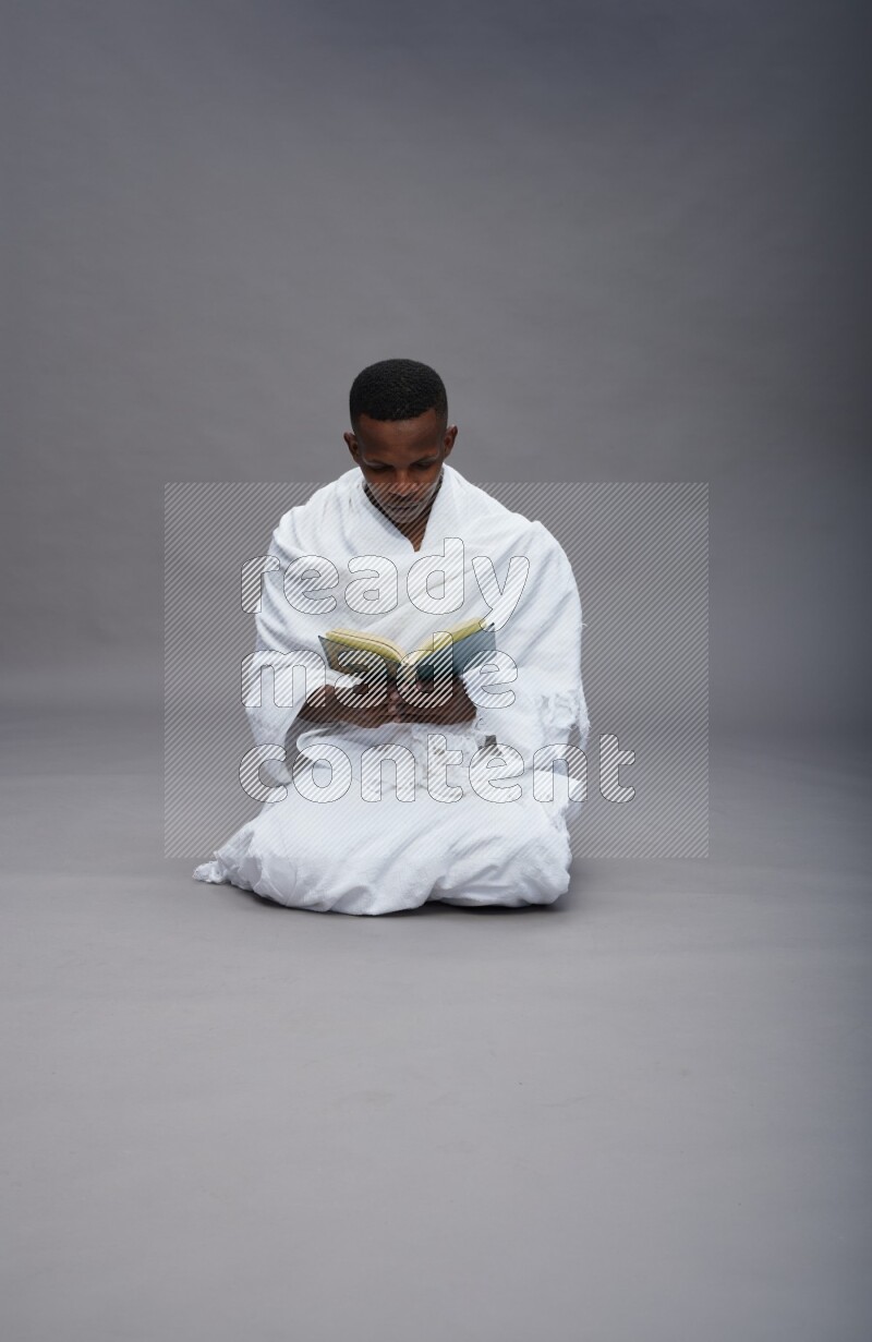 A man wearing Ehram sitting on floor reading quran on gray background