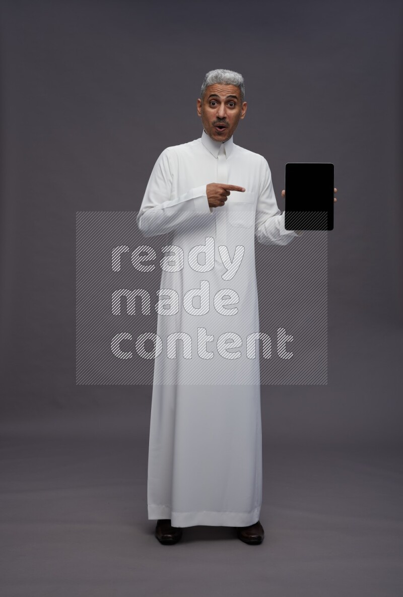 Saudi man wearing thob standing showing tablet to camera on gray background
