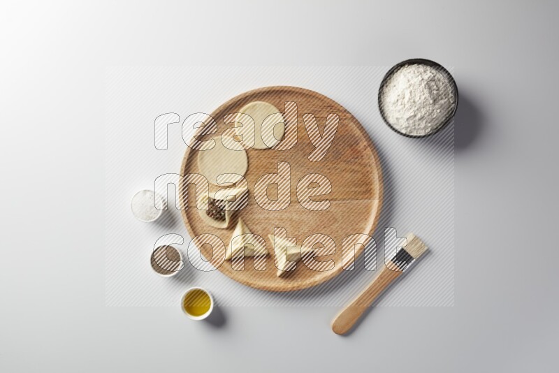 two closed sambosas and one open sambosa filled with meat while flour, salt, black pepper and oil with oil brush aside in a wooden dish on a white background