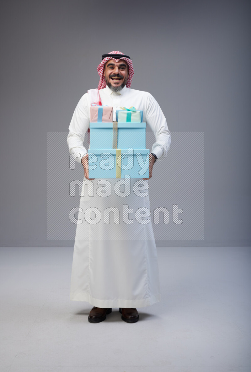 Saudi man Wearing Thob and red Shomag standing holding gift box on Gray background