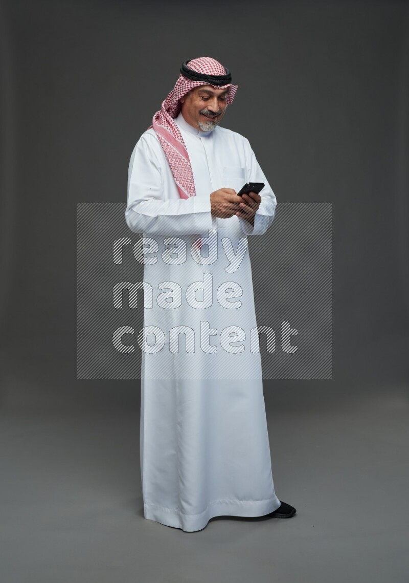Saudi man with shomag Standing texting on phone on gray background