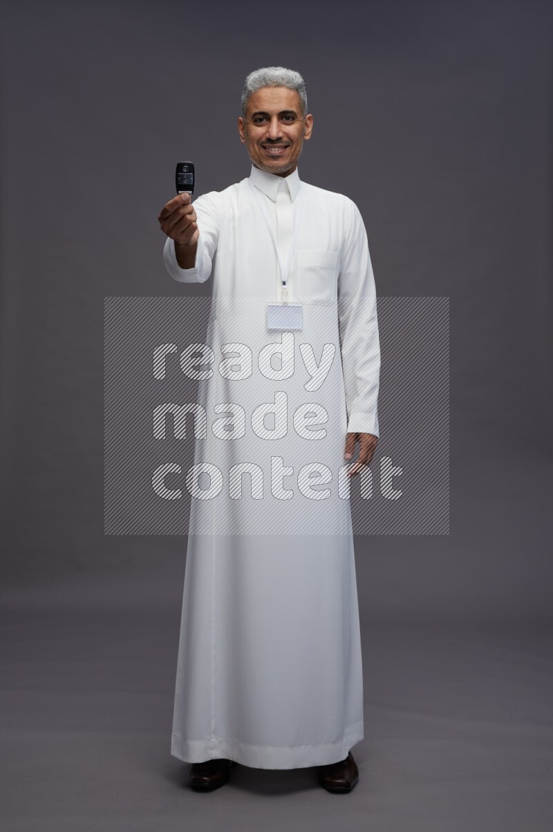 Saudi man wearing thob with neck strap employee badge standing holding key car on gray background