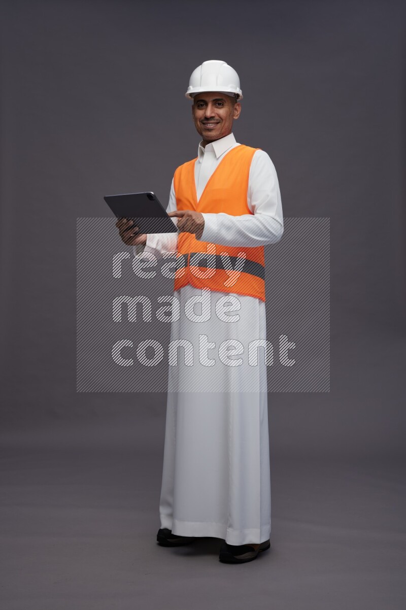 Saudi man wearing thob with engineer vest standing working on tablet on gray background