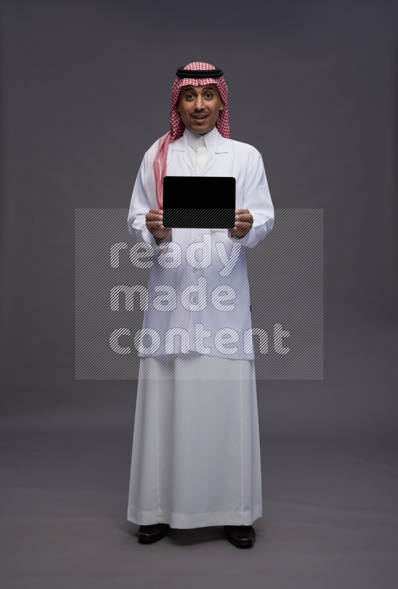 Saudi man wearing thob with lab coat and shomag with pocket employee badge standing showing tablet to camera on gray background