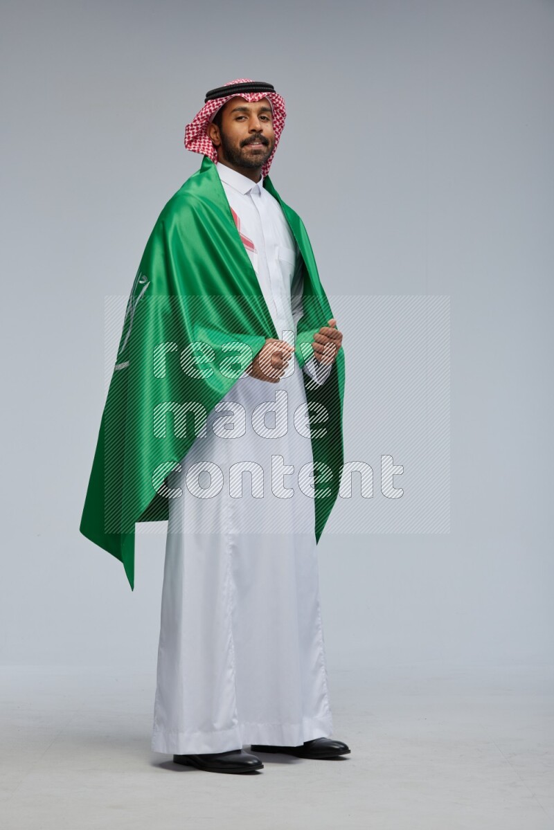 Saudi man Wearing Thob and shomag standing holding Saudi flag on Gray background