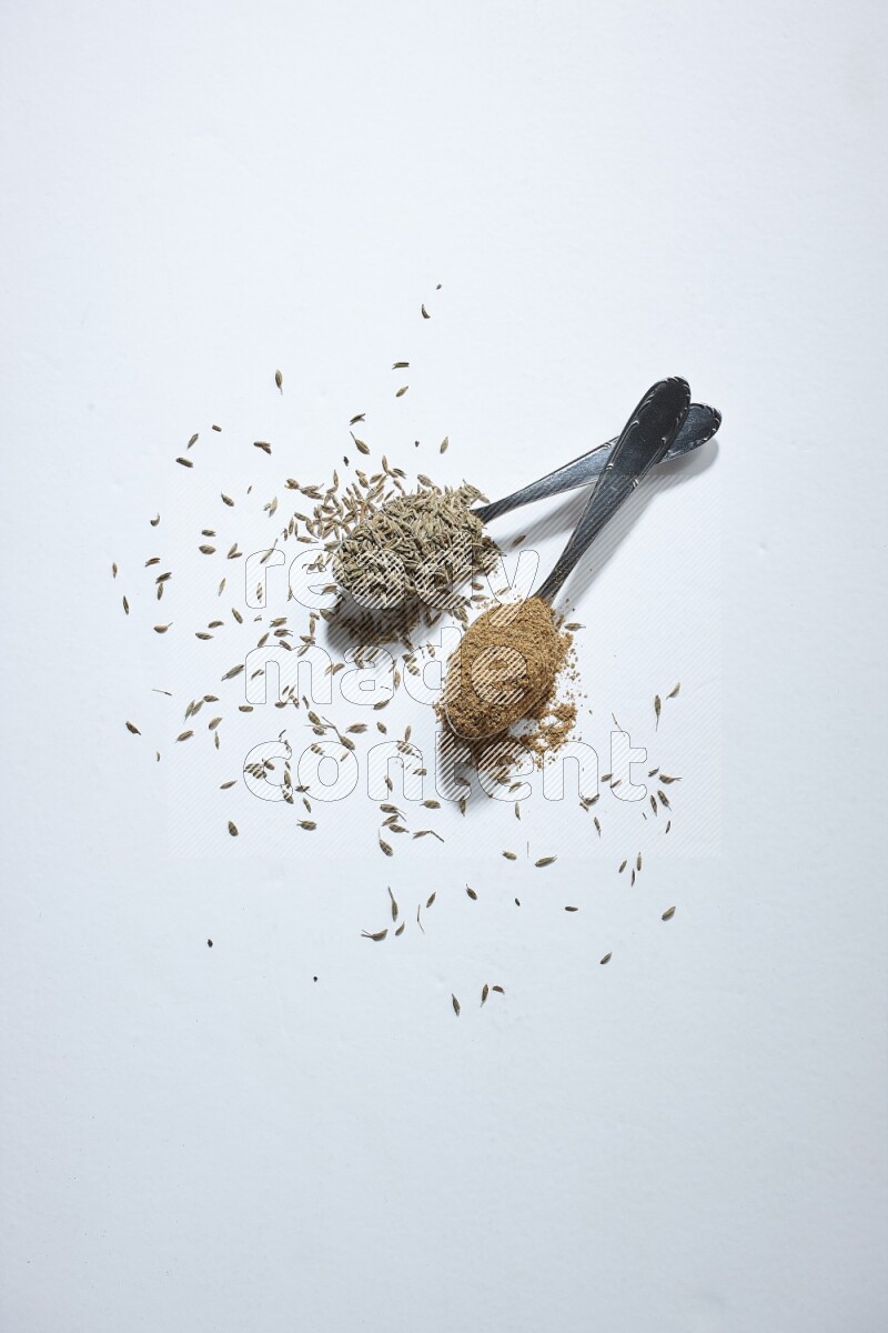 2 metal spoons full of cumin seeds and powder on white flooring
