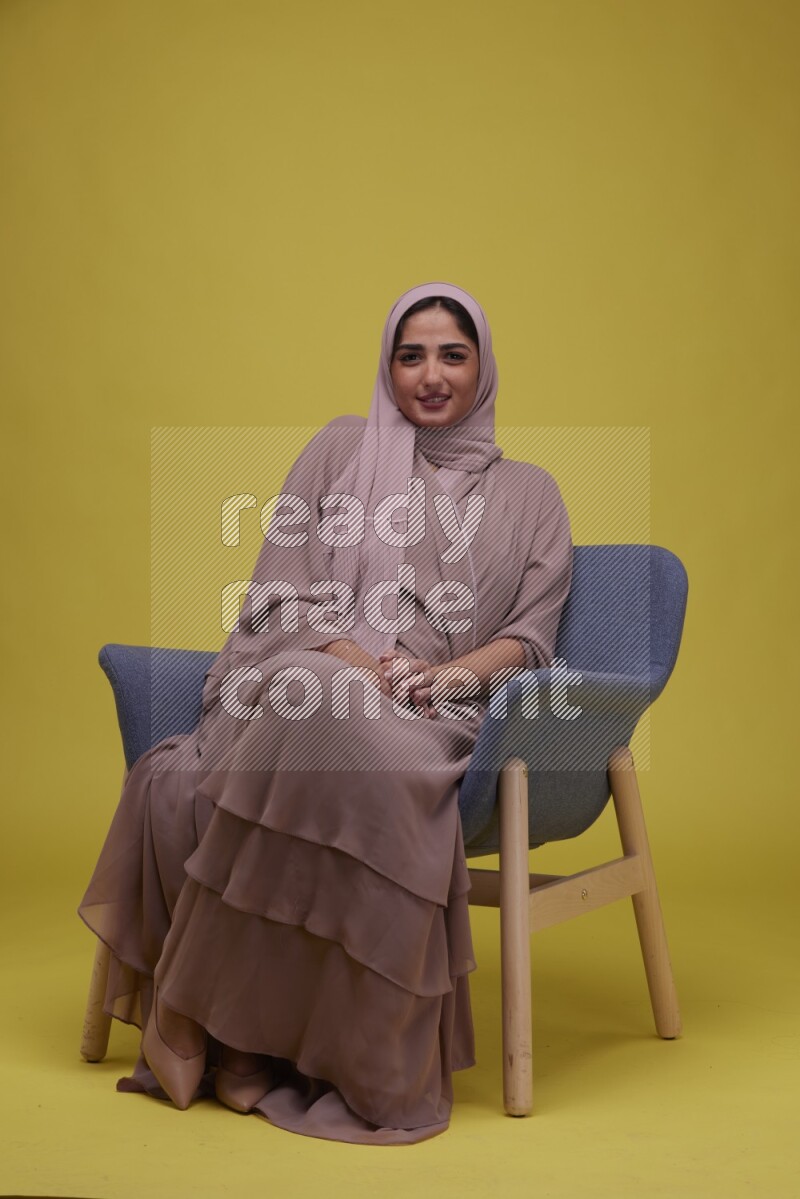 A Female sitting on a Blue Chair on a Yellow Background wearing Brown Abaya with Hijab