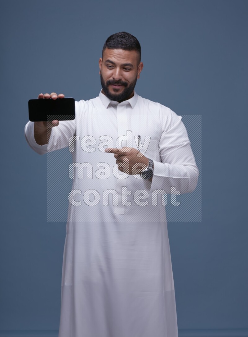 A man Showing his phone in Blue Background wearing Saudi Thob